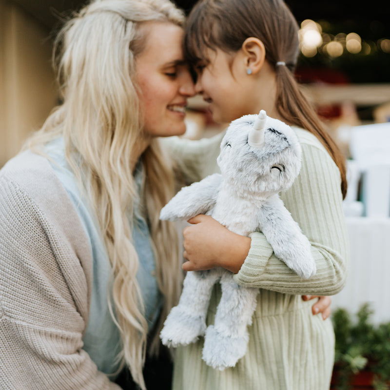 Adult woman and young girl forehead-to-forehead hugging while the girl holds a light-gray plush narwhal in a softly lit outdoor holiday setting