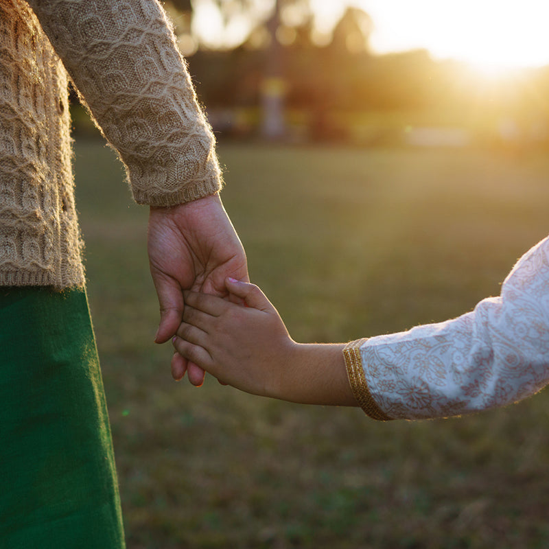 Adult holding a child’s hand in a sunlit grassy field at sunset