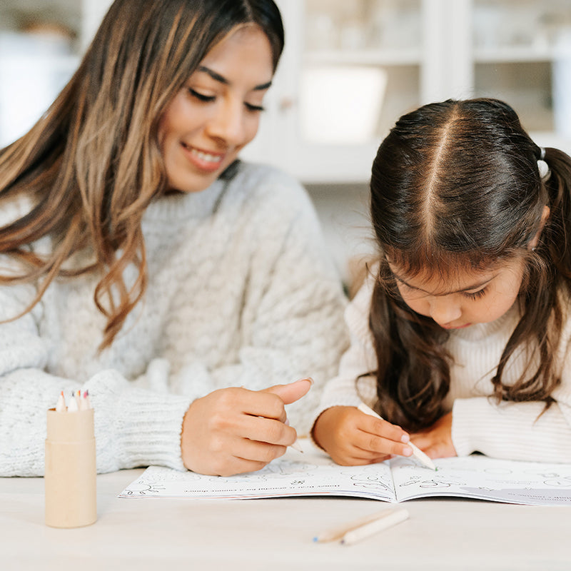 Caregiver helping a young girl color in an activity book at a kitchen table, warm cozy home setting