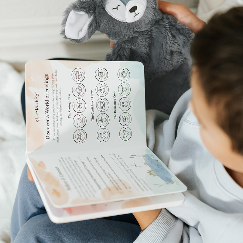 Child reading a Slumberkins board book titled Discover a World of Feelings while holding a gray plush sloth on a bed