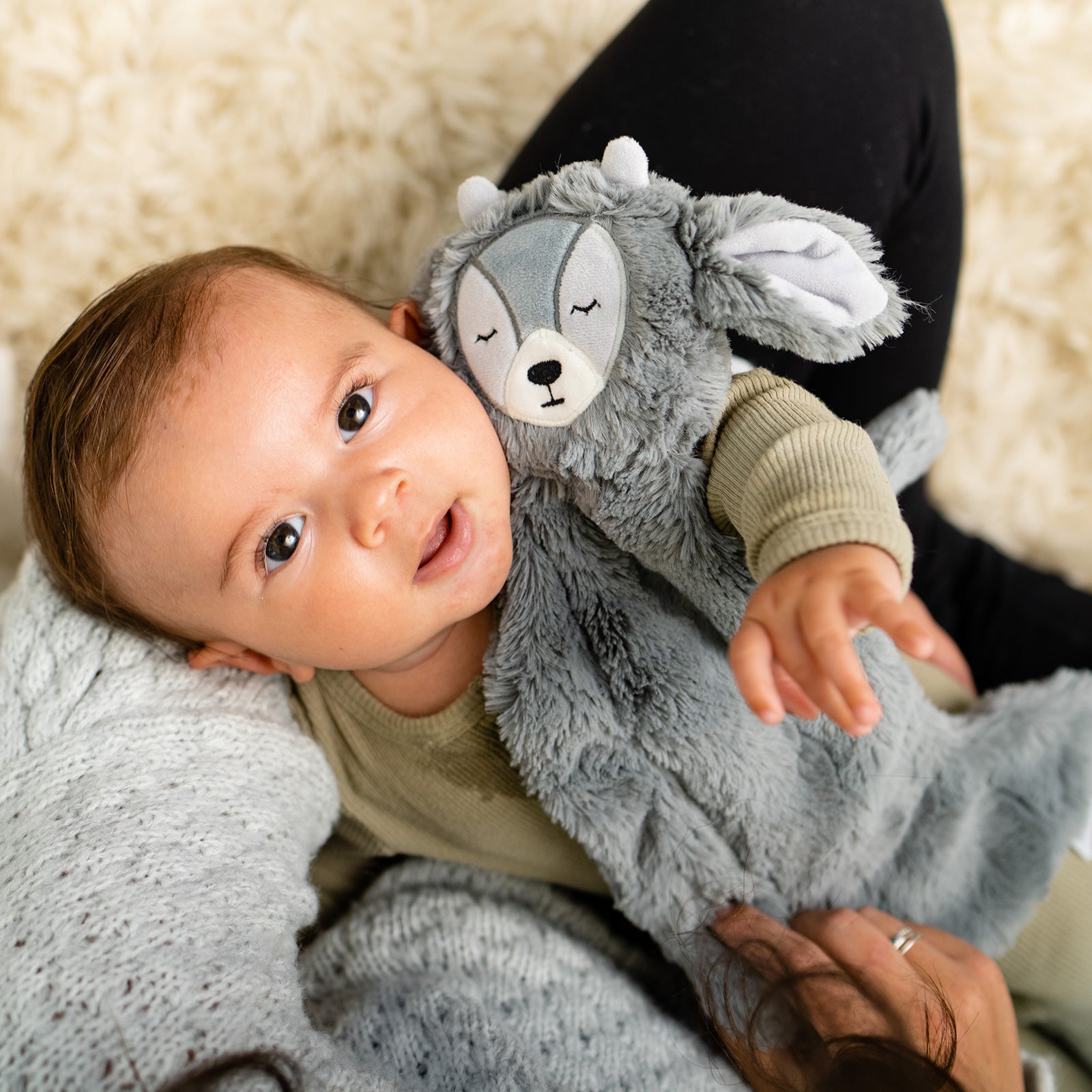 Baby holding a gray plush deer lovey while lying on a caregiver’s lap on a cream rug