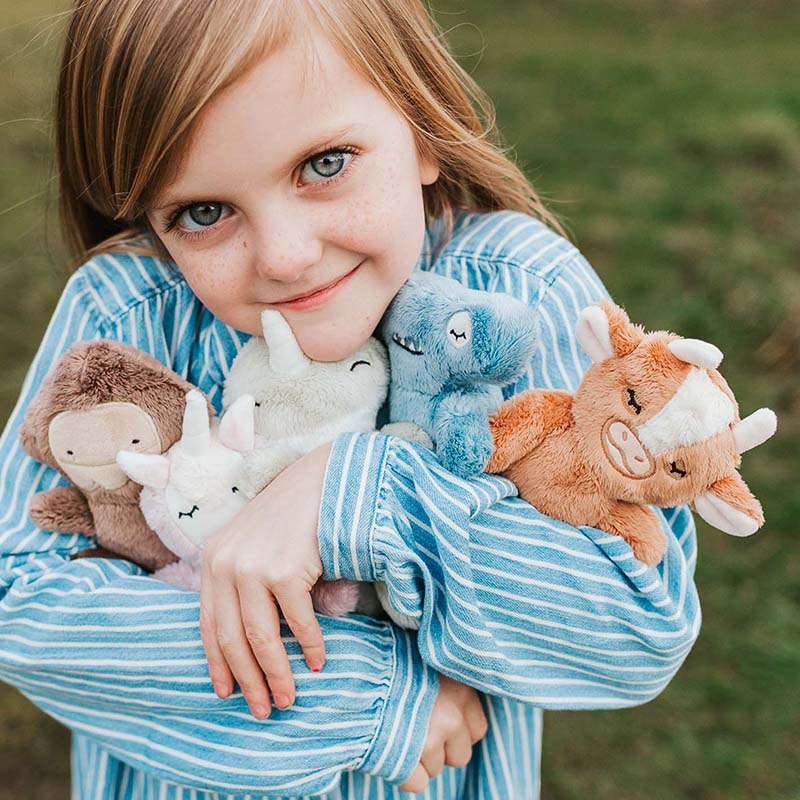 Young girl hugging five Slumberkins plush characters outdoors, smiling and wearing a blue striped shirt