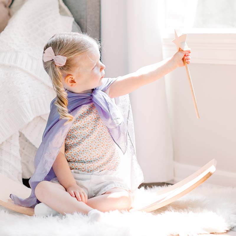 Little girl wearing a lavender cape reaches toward a star wand while sitting on a wooden rocker in a sunlit bedroom