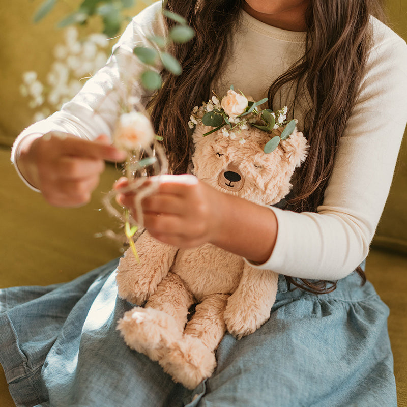 Child holding a Honey Bear plush with a flower crown, arranging small flowers while seated on a green couch