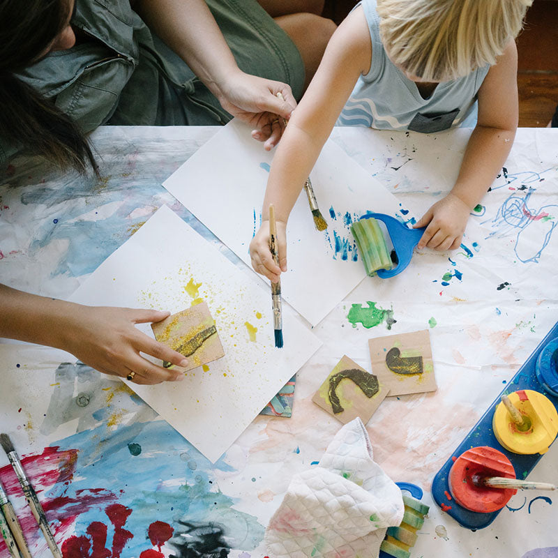 Adult and young child painting and stamping on paper at a paint-splattered table, brushes, rollers, and pots of paint visible