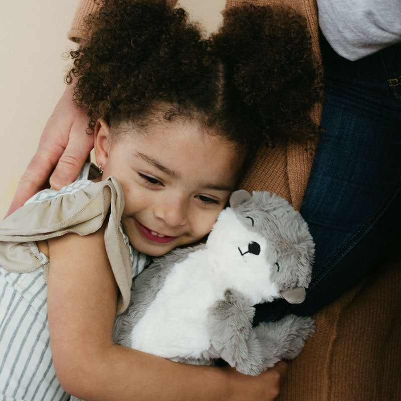Girl hugging a gray-and-white plush otter while an adult’s hand rests on her shoulder, showing comfort and connection
