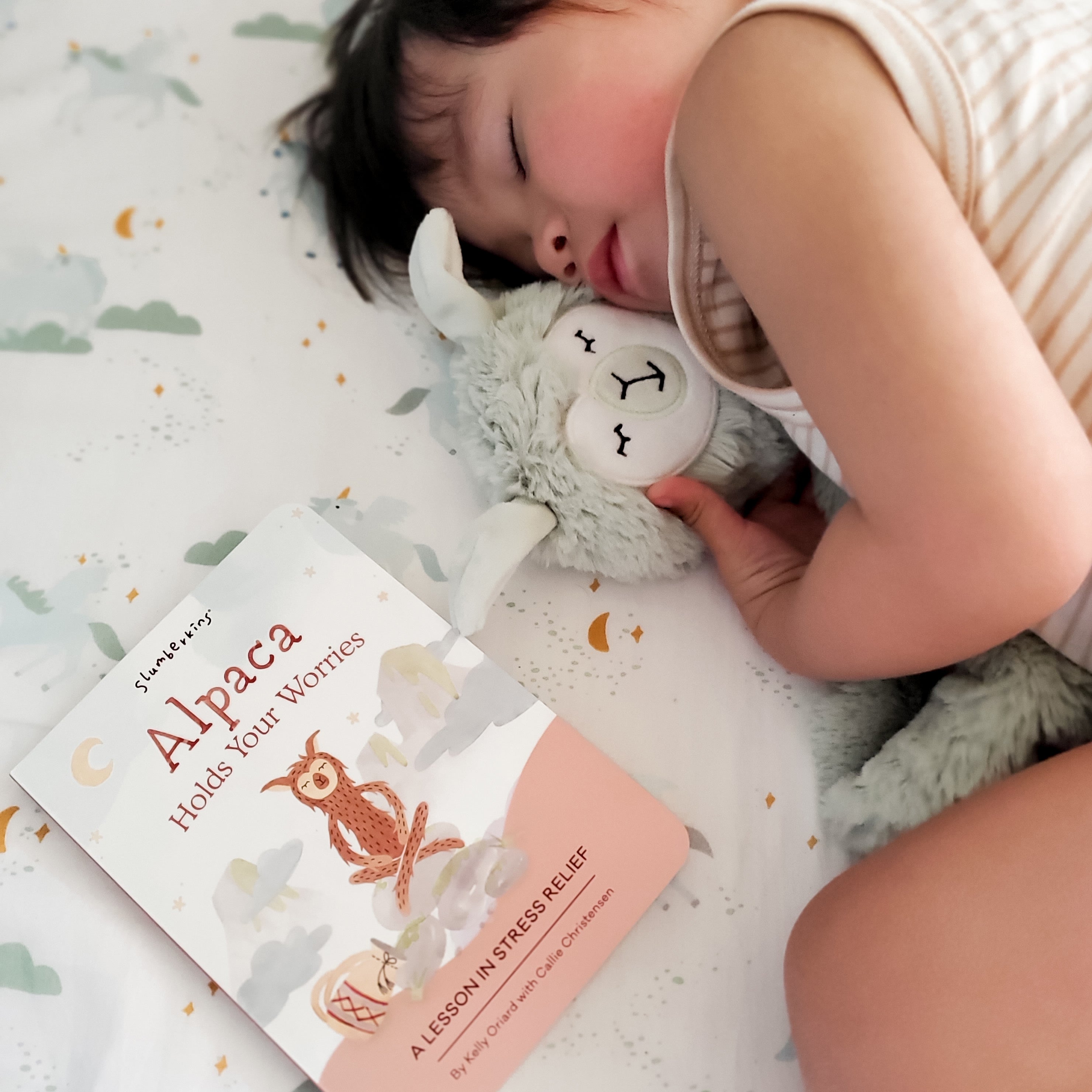 Child sleeping while hugging a plush alpaca next to a Slumberkins book on a cloud-patterned sheet