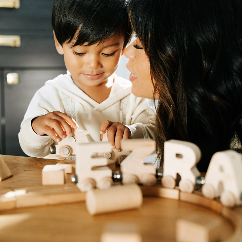 Young child playing with a wooden toy train while a caregiver watches closely at a sunlit table