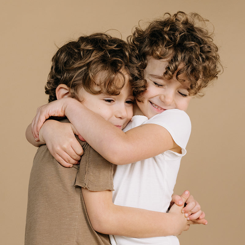 Two young children hugging and smiling, one in a white shirt and one in a brown shirt, against a beige background