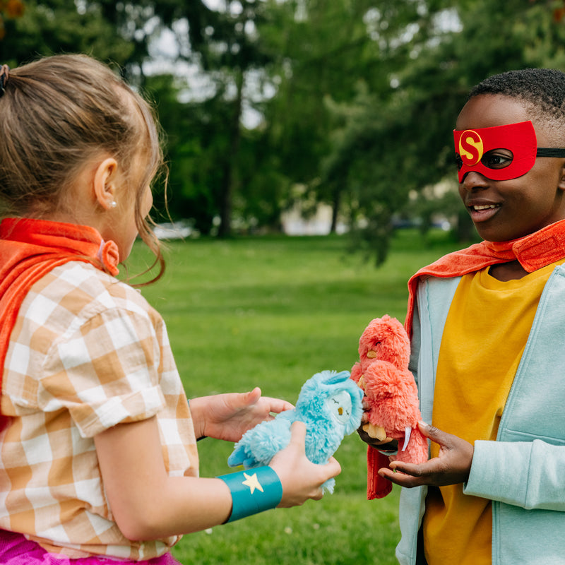 Two children wearing superhero capes exchanging plush toys in a grassy park