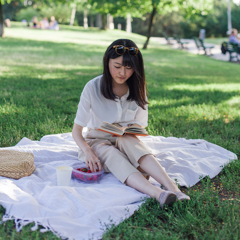 Woman sitting on a picnic blanket reading a book in a sunny park while reaching for a container of strawberries