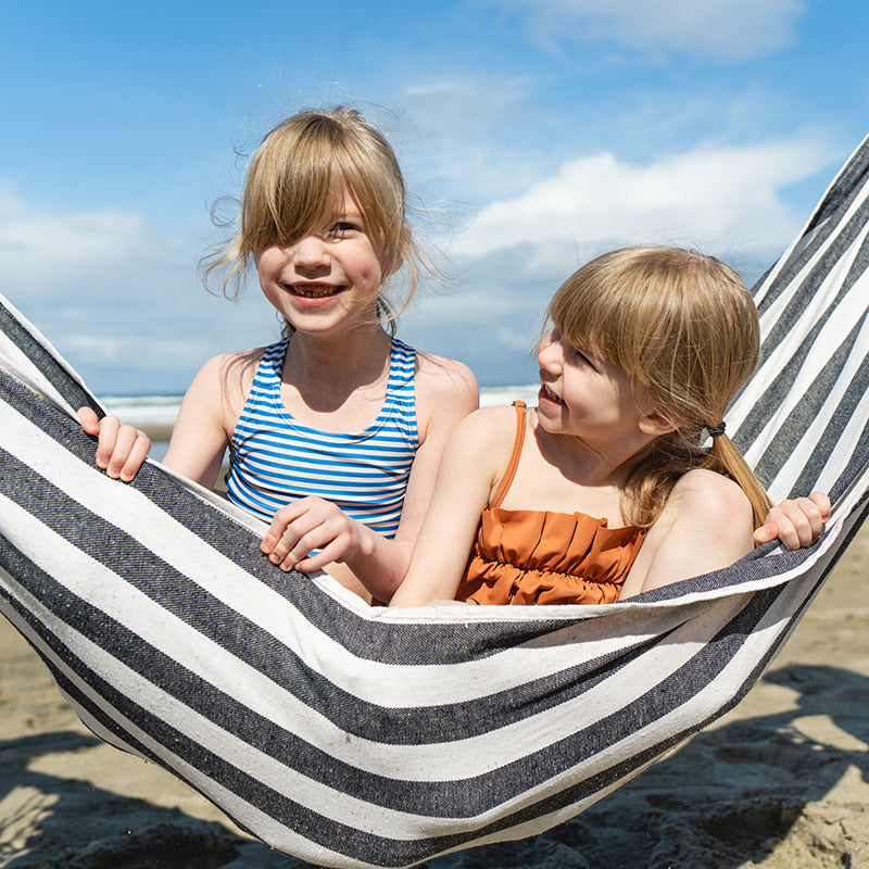 Two children sitting in a striped hammock on a sunny beach, smiling