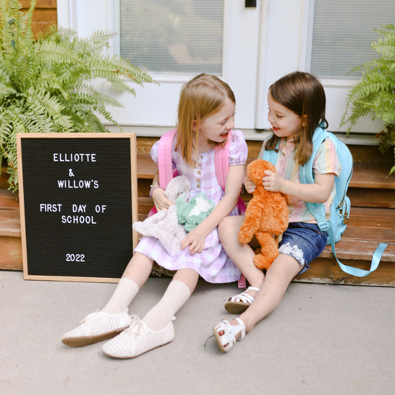 Two young girls sitting on front steps laughing with backpacks and stuffed animals beside a letter board reading Elliotte and Willow’s First Day of School 2022