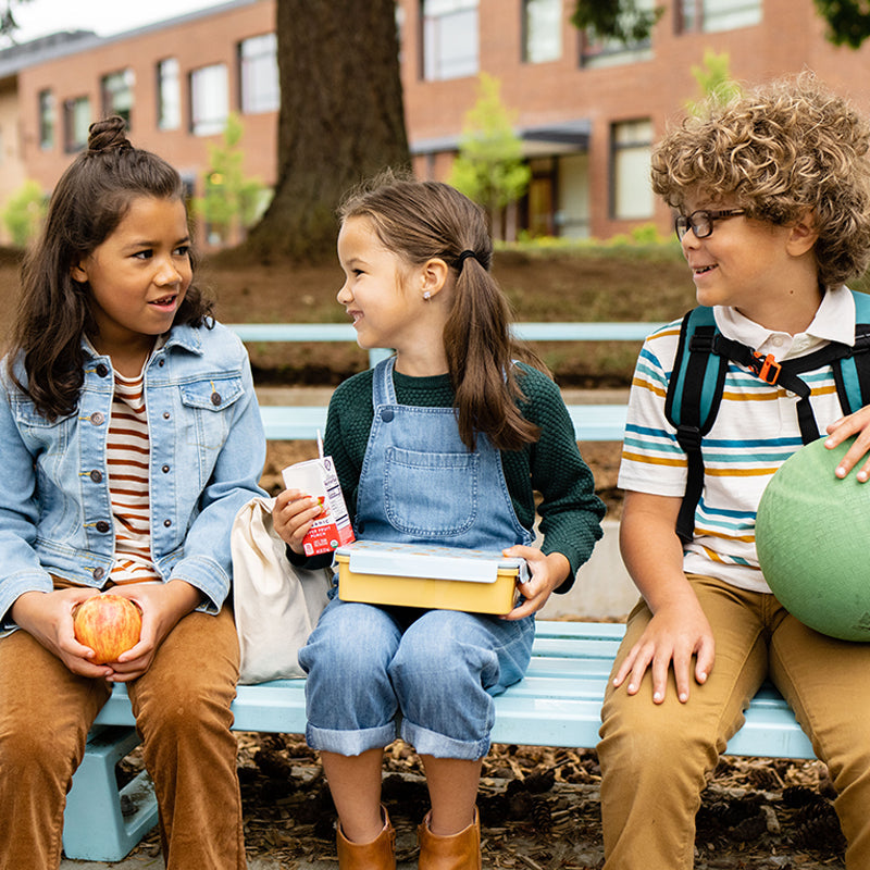 Three children sitting together on a bench outdoors, sharing a friendly moment with snacks and a ball