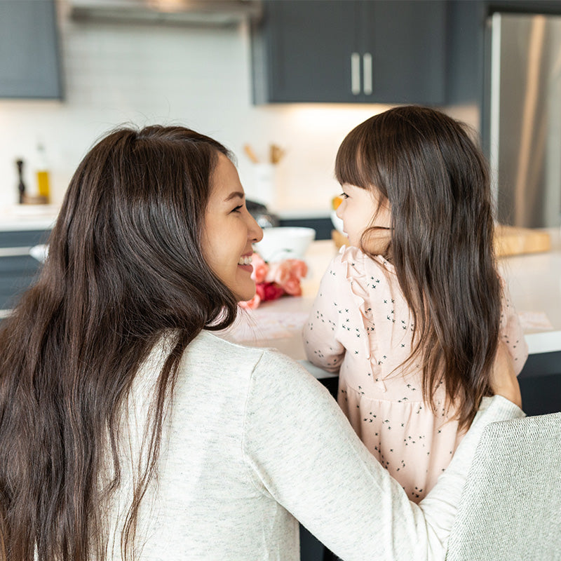 Parent smiling and hugging a young child at a kitchen counter in a bright home setting