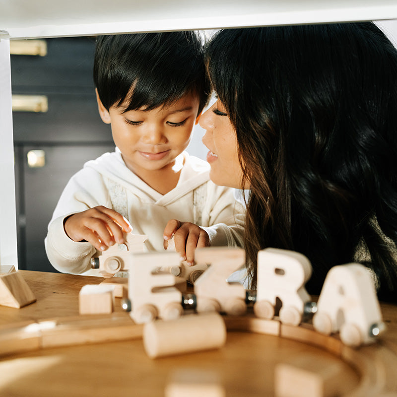 Parent leaning in while child plays with a wooden toy train on a table in a warm indoor setting