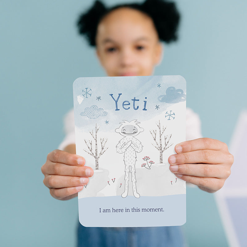Child holding a Yeti affirmation card that reads “I am here in this moment” against a soft blue background