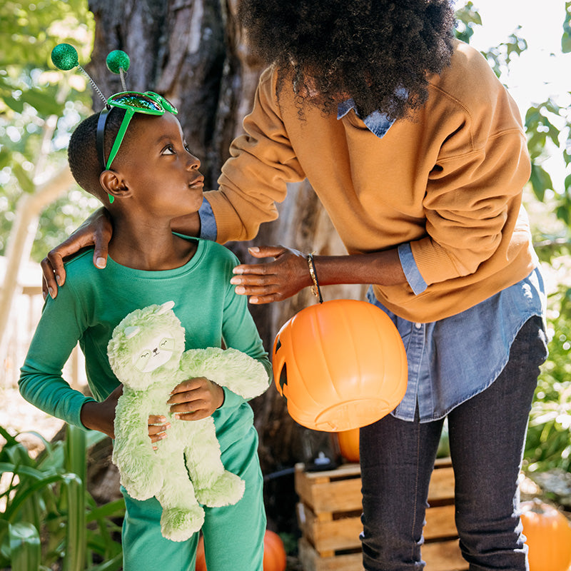 Child in green bug costume holding a green plush toy as an adult offers an orange trick-or-treat pumpkin bucket outdoors