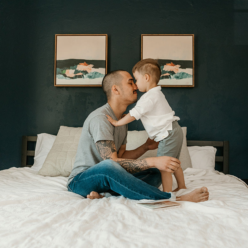 Father and toddler touching noses on a bed in a cozy bedroom with dark green wall and framed artwork