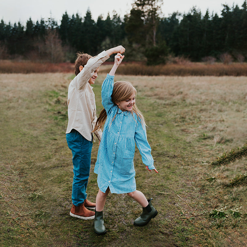 Two children twirling in a grassy field with evergreen trees in the background