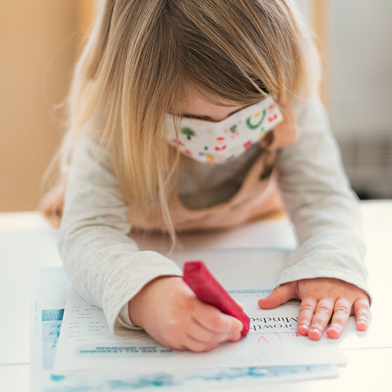 Child wearing a patterned face mask coloring a worksheet at a table, practicing social skills during COVID