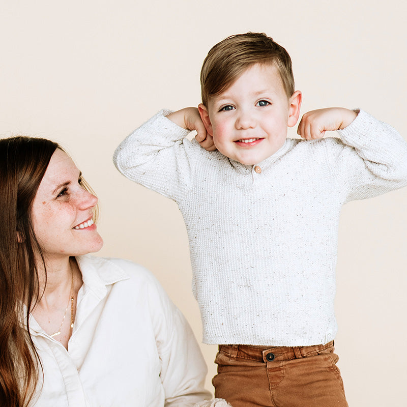 Smiling young boy flexing his arms while a woman looks up at him supportively against a neutral beige background