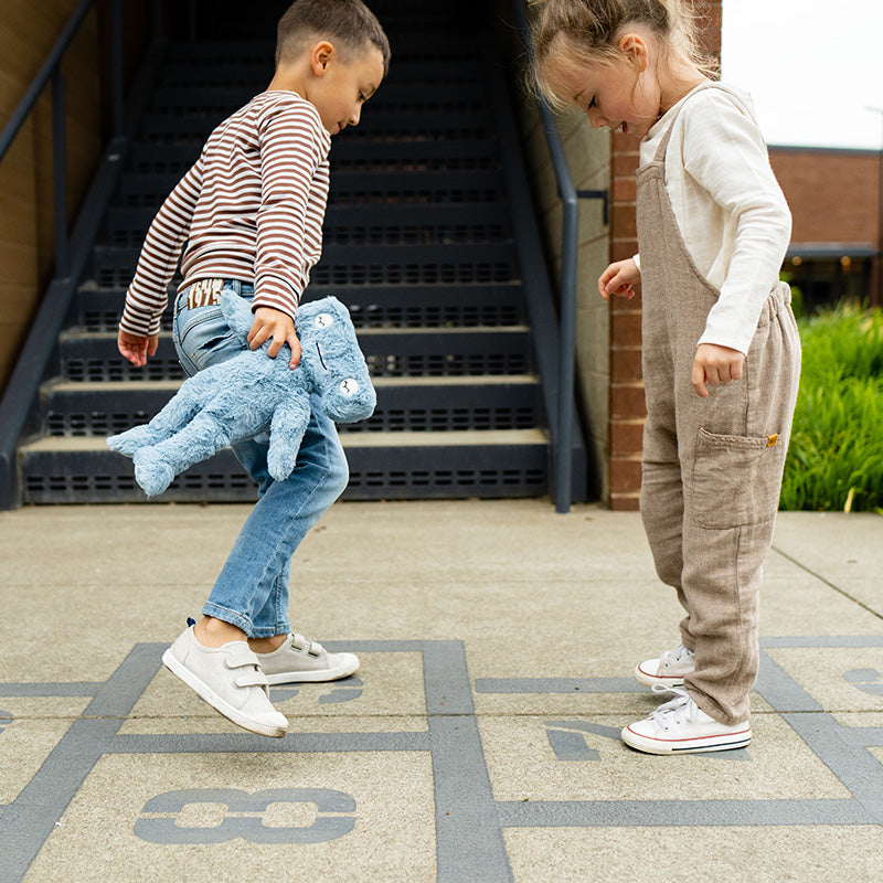 Two children playing hopscotch outside, one holding a blue plush toy; headline: 3 Social Skills for Kids and Why They’re Important