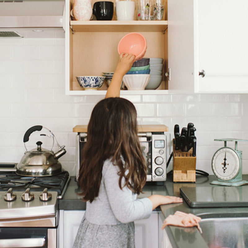 Child reaching into a kitchen cabinet to put away a stack of bowls, standing at a countertop with stove and scale visible