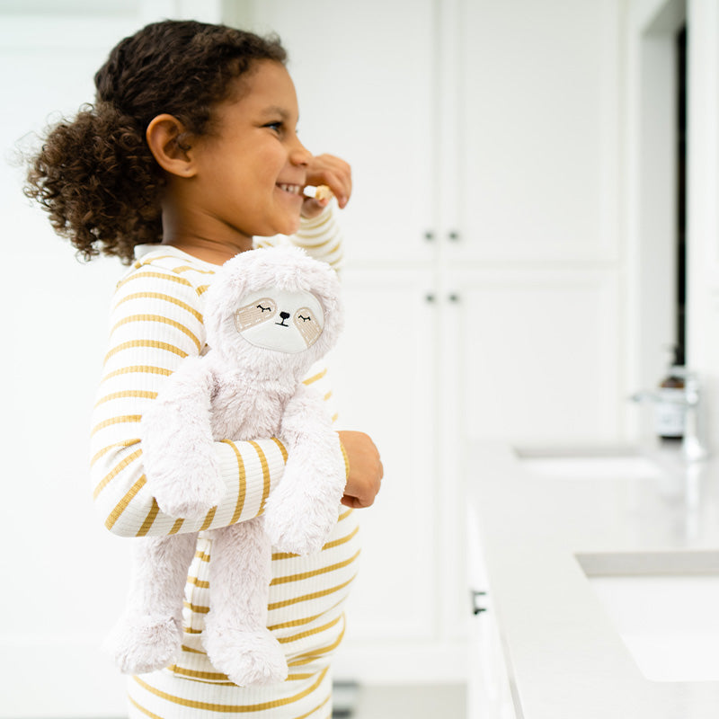 Child smiling and holding a plush sloth in a bright bathroom during a morning routine