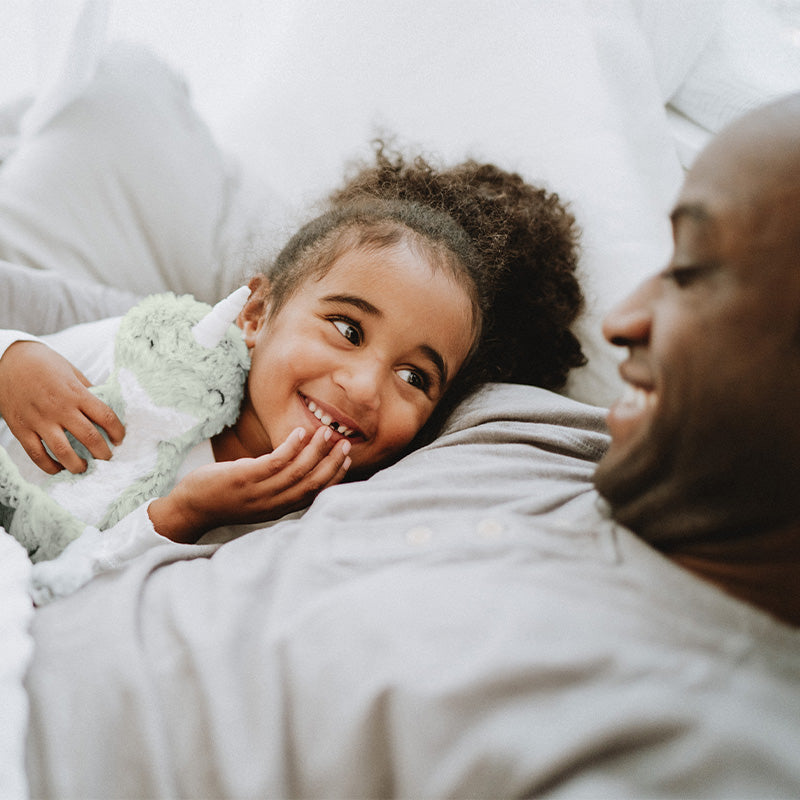 Little girl cuddling a green unicorn plush while smiling up at her father lying beside her in bed