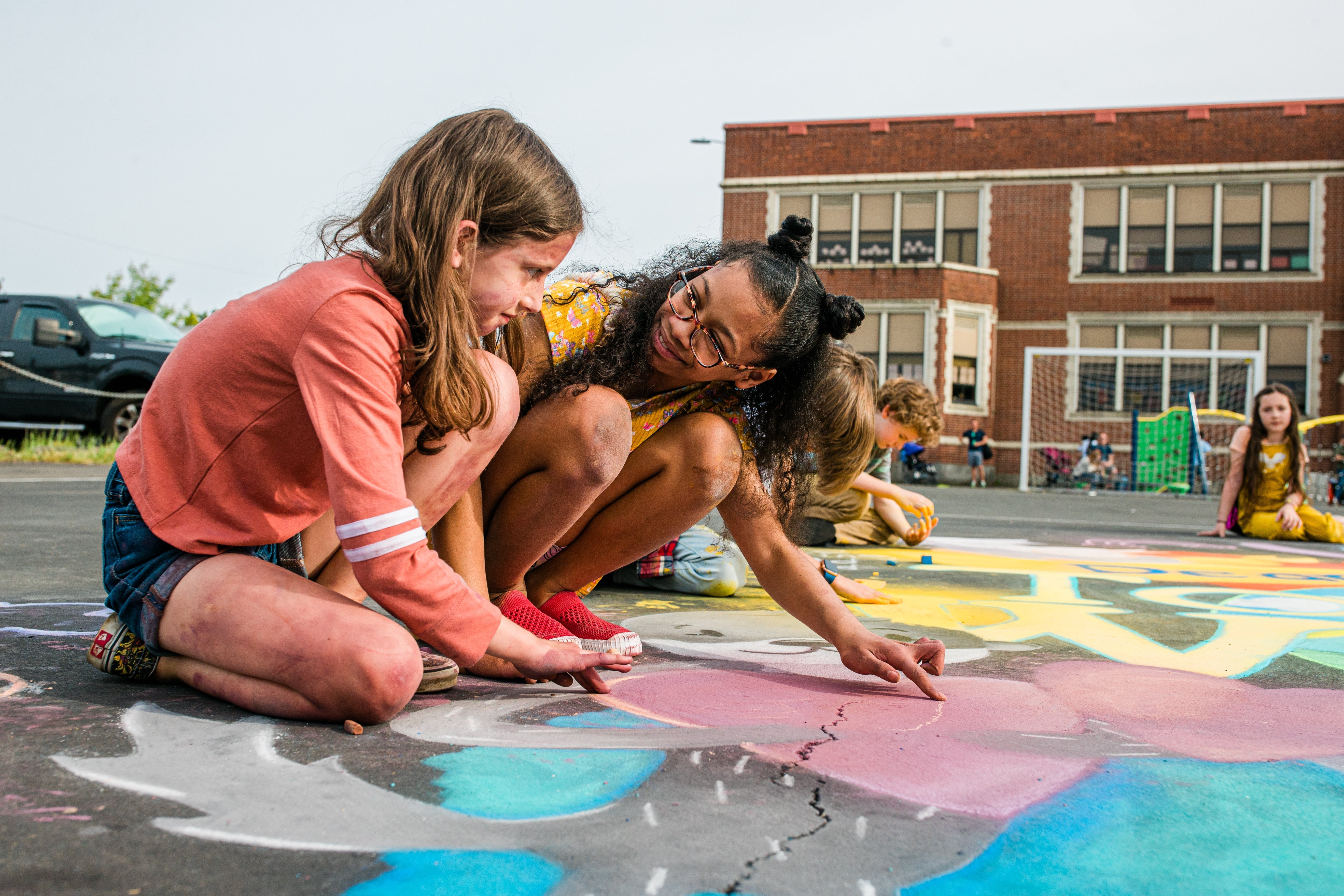 Children crouched on a school playground drawing colorful chalk art on the pavement, collaborating in front of a brick school building