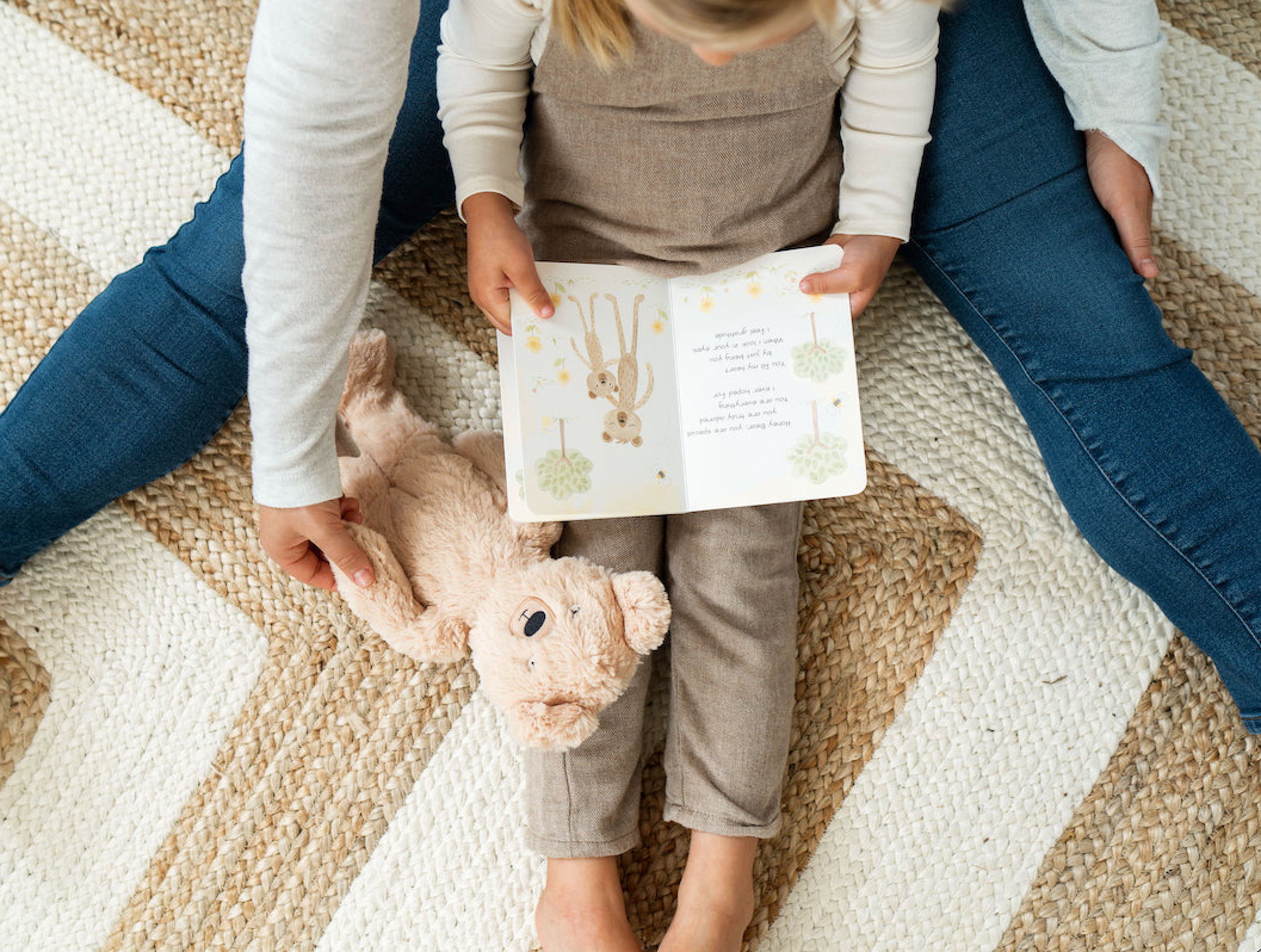 Child reading a small illustrated book on an adult’s lap with a plush bear beside them on a braided rug
