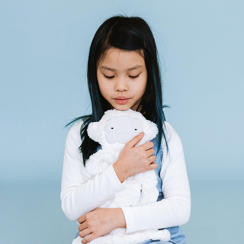 Girl hugging a white plush lamb while seated against a pale blue background, calm thoughtful expression