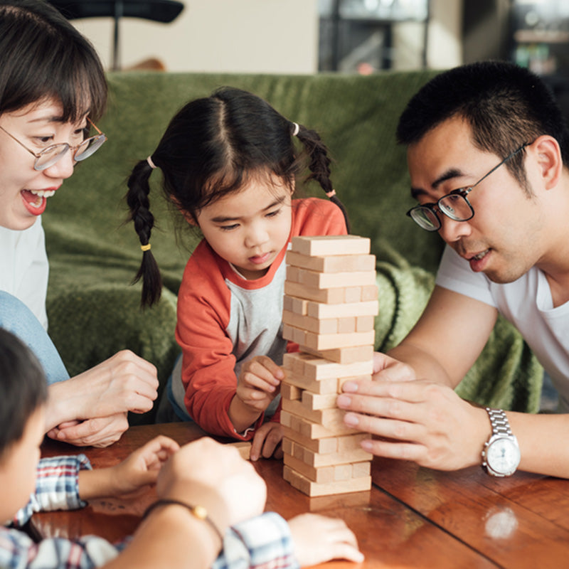 Family playing a turn-taking wooden block game at a table, parents encouraging a focused young girl