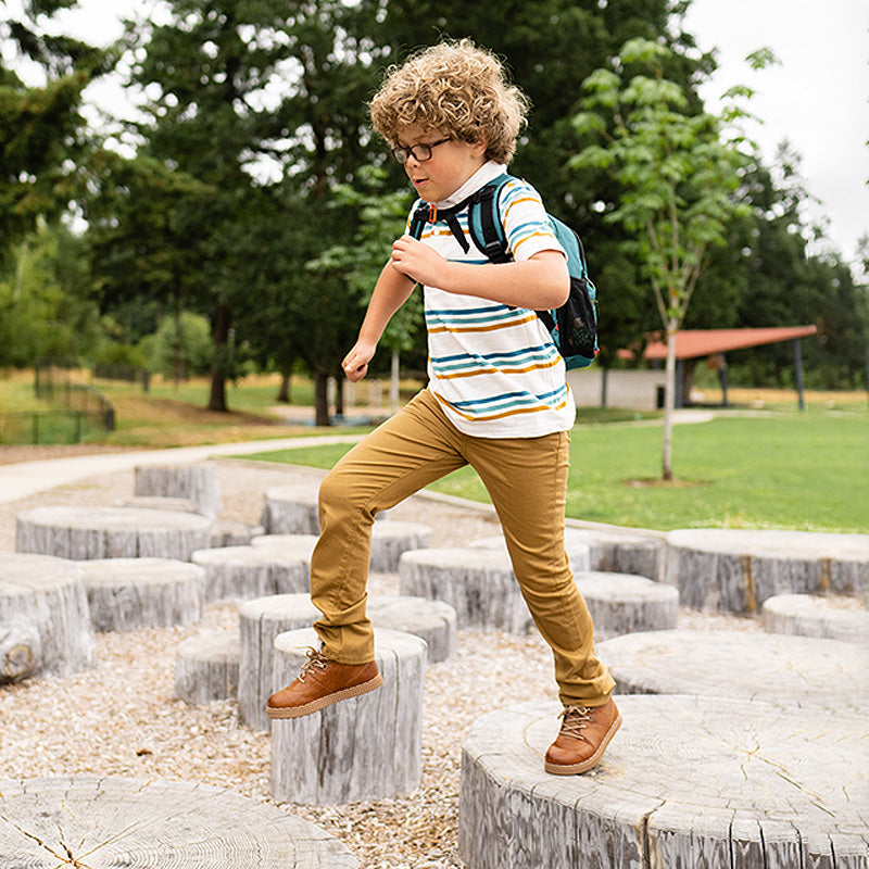 Child jumping between round wooden stepping logs at a park, wearing a striped shirt, khaki pants, and a backpack