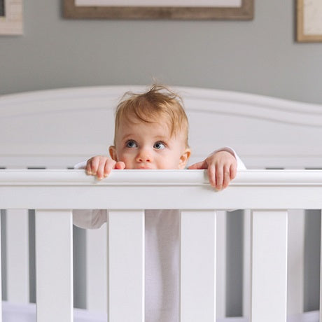 Toddler standing in a white crib peering over the railing in a softly lit nursery