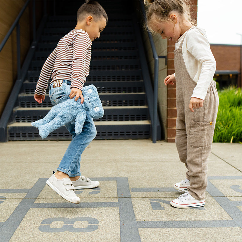 Two children playing hopscotch outside, one mid-jump holding a blue plush toy, stairs and brick building behind them