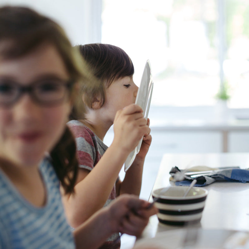 Two children at a bright kitchen table, one child licking a plate while the other smiles in the foreground