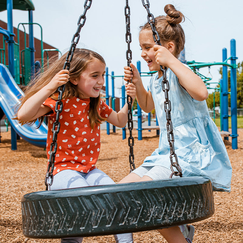Two girls laughing on a tire swing at a playground with blue play structures in the background