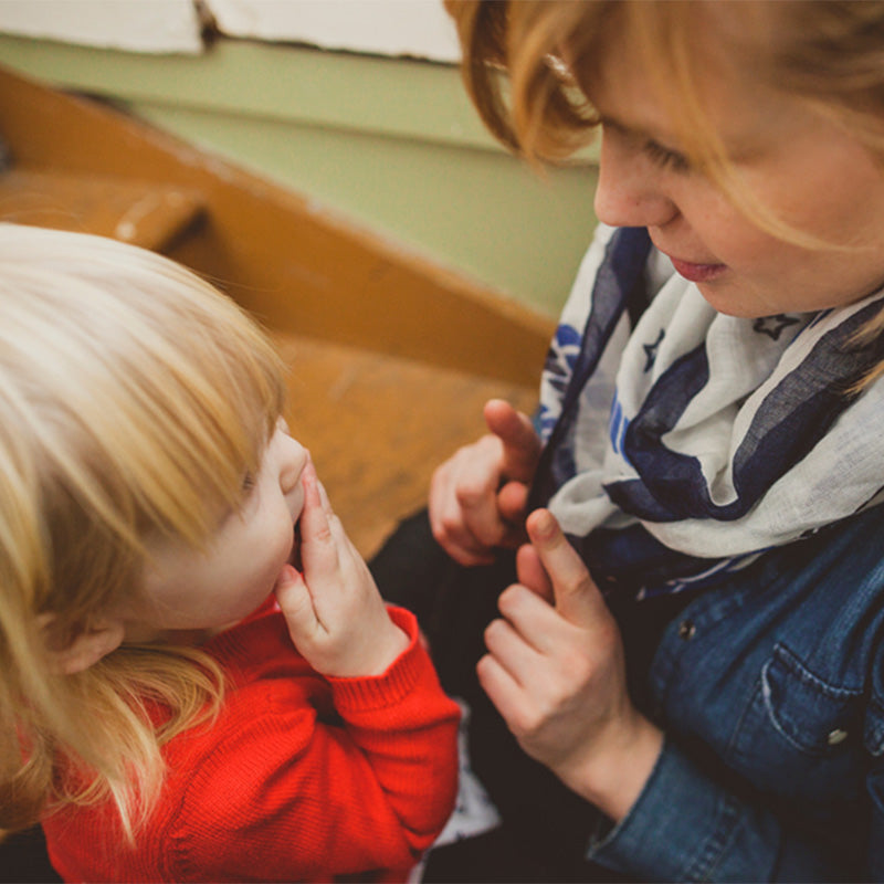 Caregiver kneeling and gently talking to a toddler who covers their mouth, close-up in a warm indoor home setting