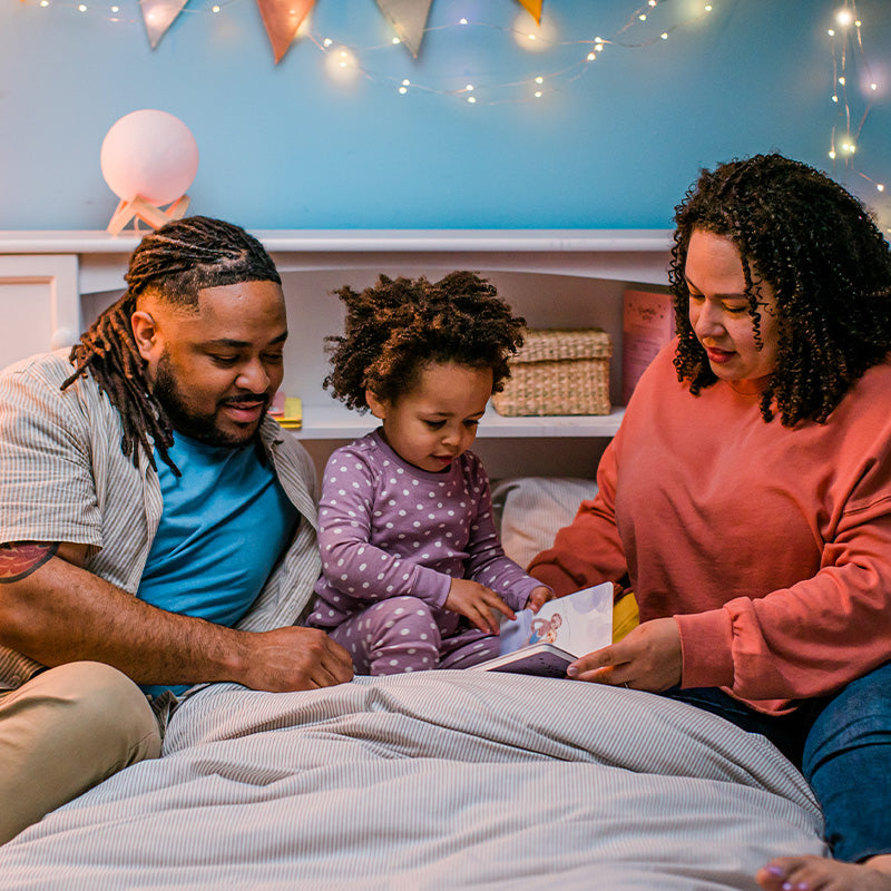 Parents reading a book with their toddler on a cozy bed under soft string lights