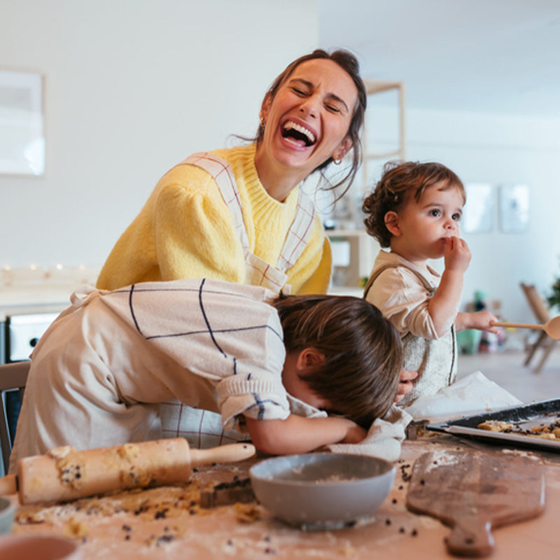Mother laughing with two young children baking at a messy kitchen table, rolling pin and mixing bowl visible