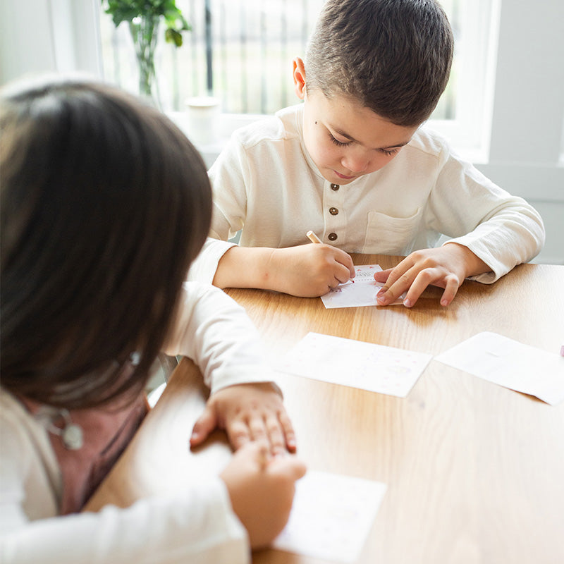 Young boy writing on a small card while a girl watches at a wooden table by a window