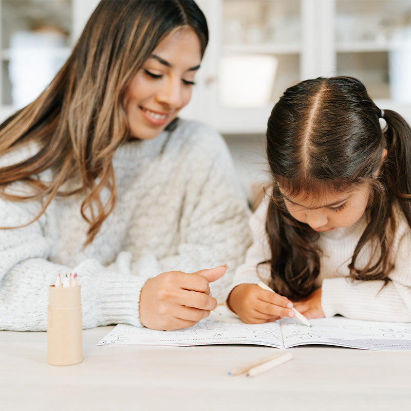 Woman helping a young girl color in a coloring book at a bright kitchen table