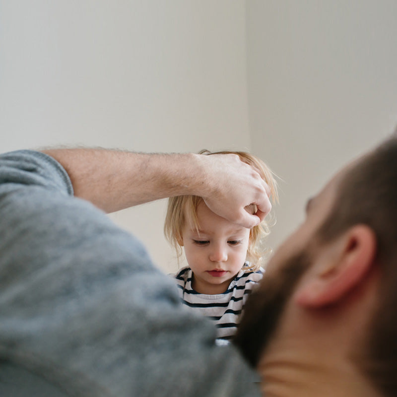 Young child looks down between two adults indoors as one rests a hand on their head