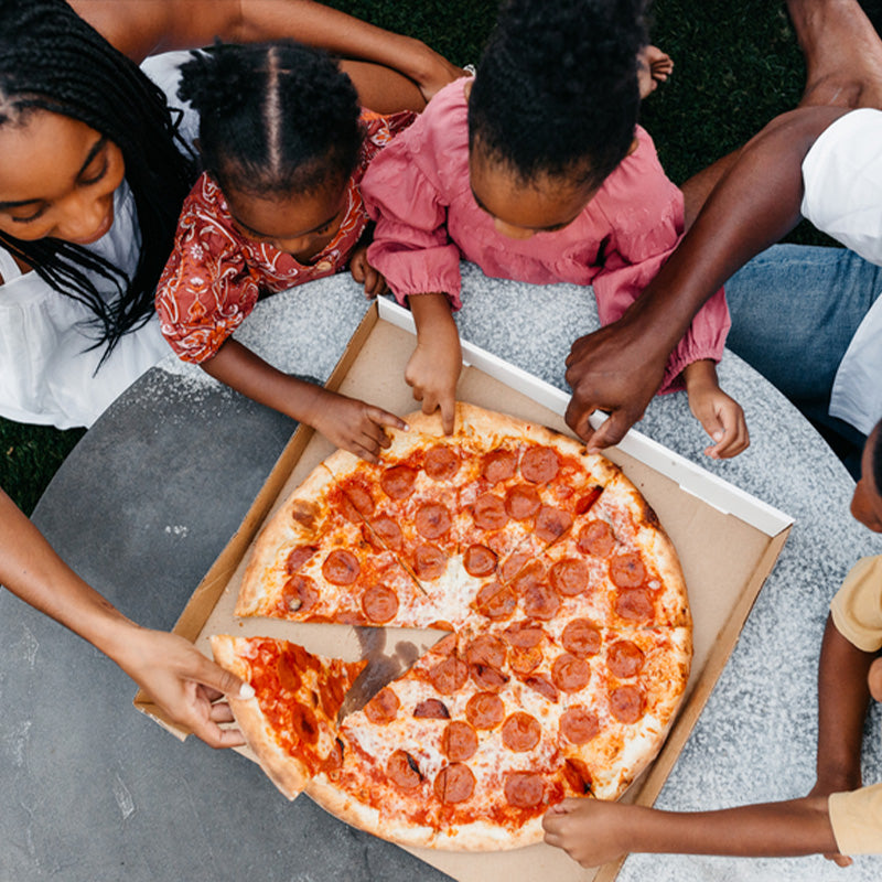 Overhead view of a family sharing a pepperoni pizza at an outdoor table, parents and children reaching for slices