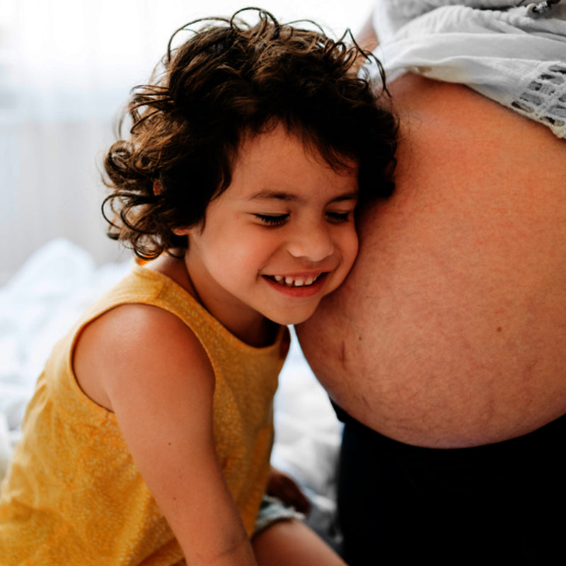Young child smiling and leaning their head against a pregnant parent’s bare belly in a sunlit bedroom