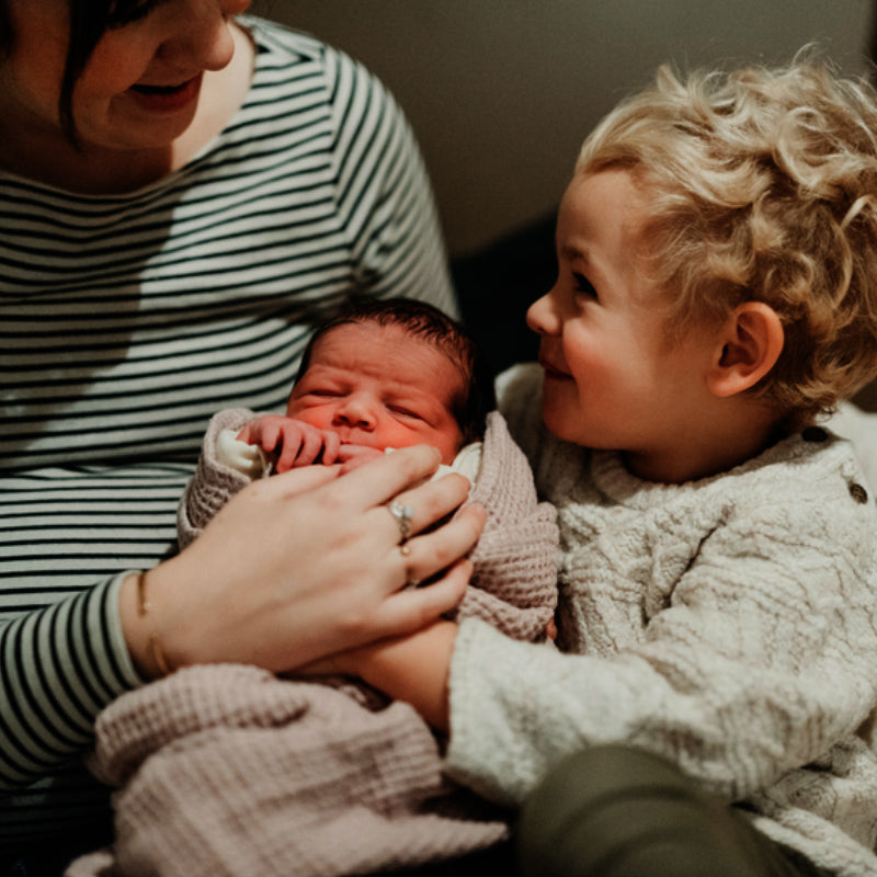 Mother in a striped shirt cradling a newborn while a smiling toddler looks on closely