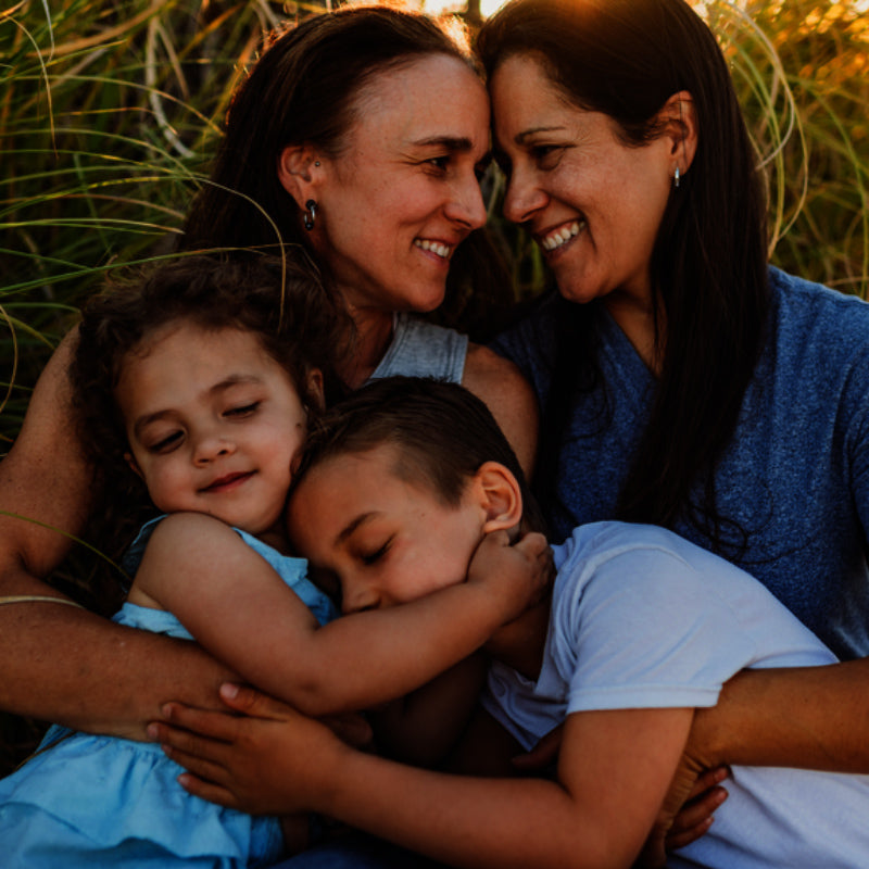 Two women smiling and embracing while holding two young children outdoors in tall grass