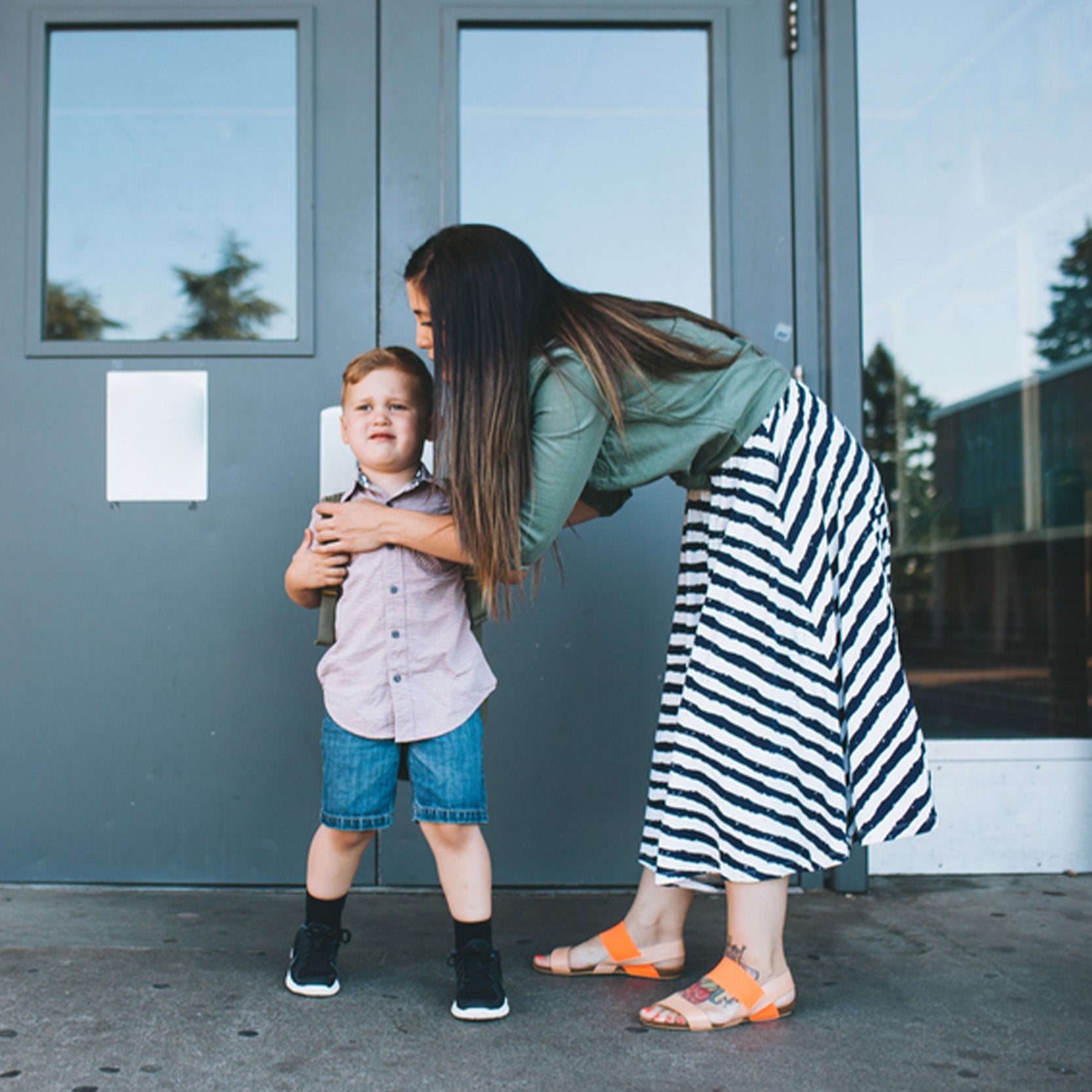 Woman bending to comfort a nervous young boy at a school or daycare doorway before drop-off
